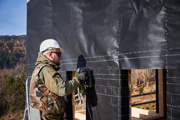 Male builder installing waterproof membrane on the wall of future cottage. Man worker building wooden frame house. Carpentry and construction concept.